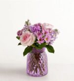 A beautiful floral arrangement featuring pink roses, purple carnations, and daisies in a textured purple glass vase.