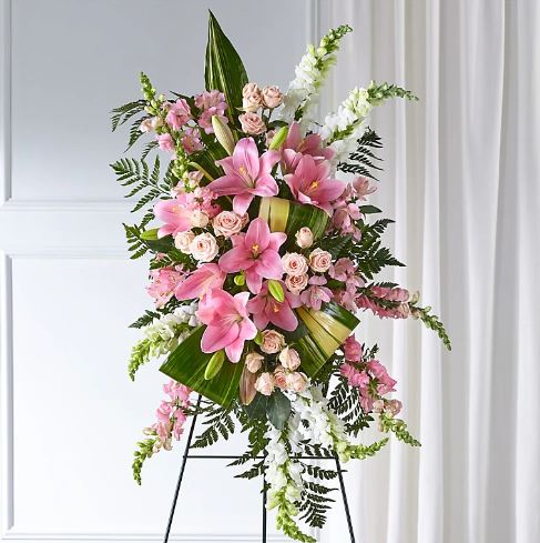 A vertical standing floral spray featuring pink lilies, peach spray roses, white and pink snapdragons, and lush greenery on a wire easel.