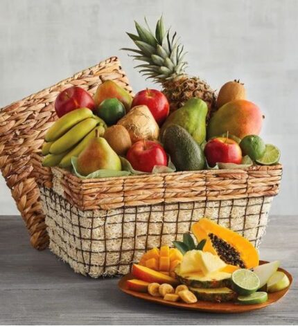 A large woven wicker basket filled with a variety of fresh whole fruits including a pineapple, apples, pears, bananas, and kiwi, with a small wooden plate in the foreground displaying sliced tropical fruits.