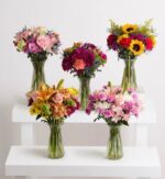 Five vibrant floral arrangements in clear glass vases, displayed on tiered white rectangular pedestals against a plain off-white background. The bouquets feature a variety of flowers including sunflowers, roses, lilies, and carnations in diverse colors.