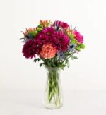A vibrant "Fresh Market Bouquet Deluxe" featuring magenta chrysanthemums, peach carnations, blue sea holly (eryngium), and green button poms in a clear fluted glass vase.