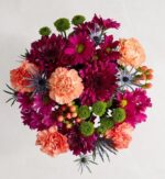 A vibrant top-down view of a "Fresh Market Deluxe" floral bouquet featuring a mix of deep magenta chrysanthemums, peach-colored carnations, green button pompons, blue sea holly (eryngium), and hypericum berries against a plain light grey background.