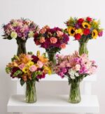 Five diverse floral bouquets in glass vases arranged on white tiered pedestals against a neutral background.