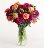 A vibrant premium floral arrangement in a clear glass vase featuring peach carnations, magenta chrysanthemums, blue sea holly (eryngium), and green button poms.