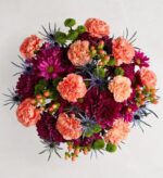 A vibrant top-down view of a dense floral bouquet featuring a rich mix of peach-colored carnations, deep magenta chrysanthemums, blue sea holly (eryngium), and green button poms accented with pink hypericum berries.