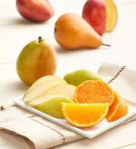 A close-up high-angle shot of fresh autumnal fruits featuring whole and sliced pears, a red apple, and vibrant orange slices arranged on a white rectangular plate and a light wooden surface.