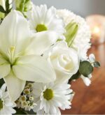 A close-up photograph of a "Healing Tears" all-white floral arrangement featuring a large lily, a white rose, white daisies with yellow centers, and green eucalyptus leaves against a soft-focus background with a candle.