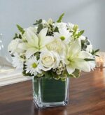 An elegant "Healing Tears" all-white floral arrangement featuring white lilies, roses, and daisies in a square glass vase lined with green leaves, set on a wooden surface.