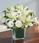 A premium "Healing Tears" all-white floral arrangement featuring white lilies, roses, carnations, and snapdragons in a square glass vase lined with green leaf wrap, set on a wooden table.
