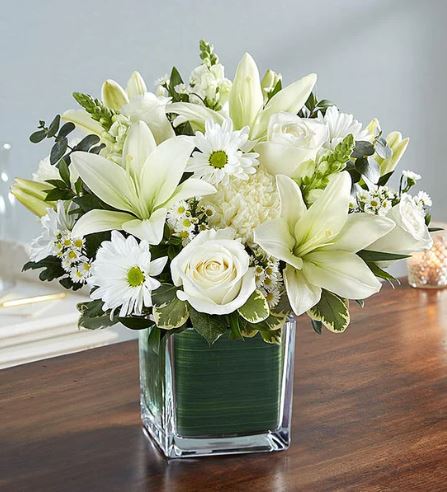 A premium "Healing Tears" all-white floral arrangement featuring white lilies, roses, carnations, and snapdragons in a square glass vase lined with green leaf wrap, set on a wooden table.