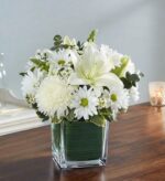 An elegant all-white floral arrangement featuring large lilies, chrysanthemums, and daisies in a glass cube vase lined with green leaves.