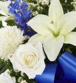 A close-up of a "Heartfelt Sympathies" funeral standing basket featuring white lilies, a white rose, white mums, blue delphiniums, and small daisies accented with a vibrant blue ribbon.