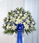 A large, fan-shaped funeral standing basket arrangement featuring white lilies, roses, and chrysanthemums accented with blue delphinium and lush greenery, finished with a prominent blue ribbon.