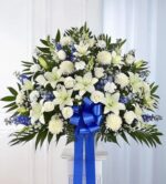 A large, fan-shaped funeral standing basket arrangement featuring white lilies, roses, and chrysanthemums accented with blue delphinium and lush greenery, finished with a large blue satin bow.