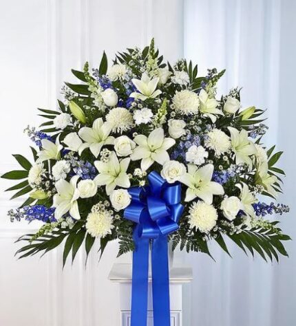 A large, fan-shaped funeral standing basket arrangement featuring white lilies, roses, and chrysanthemums accented with blue delphinium and lush greenery, finished with a large blue satin bow.