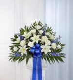 A symmetrical funeral standing basket arrangement featuring white lilies, roses, chrysanthemums, and blue delphinium, accented with lush greenery and a large blue satin ribbon.