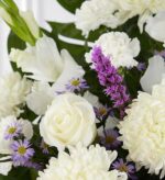 A close-up of a floral arrangement featuring white roses, white carnations, white chrysanthemums, and purple Liatris with small lavender daisies against a backdrop of green leaves.