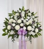 A large, fan-shaped funeral standing spray composed of white roses, white carnations, white chrysanthemums, and lavender accents, finished with a large lavender satin ribbon.