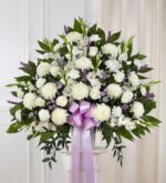 A large, premium standing funeral spray arrangement featuring a dense mix of white mums, white roses, and lavender accents, adorned with a large light purple satin ribbon.