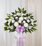 A large, fan-shaped funeral flower arrangement featuring white roses, large white chrysanthemums, and lavender accents, adorned with a prominent light purple satin ribbon.
