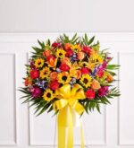 A vibrant and large sympathy standing basket floral arrangement featuring a mix of sunflowers, orange roses, red carnations, and purple filler flowers, accented with lush greenery and a large yellow ribbon.