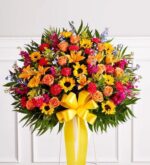A large, vibrant standing funeral spray featuring a sunburst of sunflowers, orange roses, lilies, and magenta carnations, accented with blue delphinium and a large yellow bow.