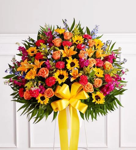 A large, vibrant standing funeral spray featuring a sunburst of sunflowers, orange roses, lilies, and magenta carnations, accented with blue delphinium and a large yellow bow.