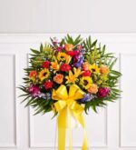 A vibrant and colorful standing floral sympathy basket featuring sunflowers, orange roses, lilies, and red carnations, accented with a large yellow bow.