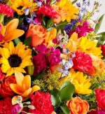 A vibrant and dense floral arrangement featuring sunflowers, orange roses, pink carnations, and blue delphiniums.