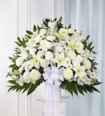 A large, elegant white funeral standing basket arrangement featuring lilies, roses, carnations, and chrysanthemums with lush greenery and a prominent white satin ribbon.