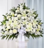 Large white funeral standing basket arrangement featuring lilies, roses, carnations, and gladioli with a white ribbon on a pedestal.