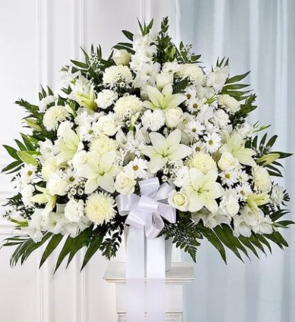 Large white funeral standing basket arrangement featuring lilies, roses, carnations, and gladioli with a white ribbon on a pedestal.