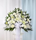 A large, fan-shaped funeral standing basket arrangement featuring an elegant mix of all-white flowers, including lilies, roses, and carnations, accented with lush greenery and a prominent white satin ribbon.