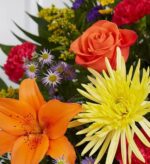 A vibrant floral arrangement featuring an orange lily, a bright yellow spider mum, an orange rose, and various purple and red accent flowers among green foliage.