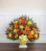 A large, fan-shaped "Heartfelt Tribute" floral floor basket featuring a vibrant mix of orange lilies, yellow chrysanthemums, red carnations, and orange roses, accented with purple statice and lush green palm fronds, finished with a large pale yellow bow.