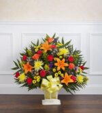 A large, fan-shaped "Heartfelt Tribute" funeral floor basket featuring orange lilies, red carnations, yellow chrysanthemums, and purple statice, accented with a large yellow bow in a white wicker basket.