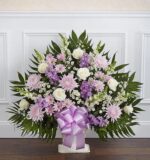 A large, fan-shaped funeral floor basket arrangement featuring white roses, lavender chrysanthemums, purple stock, and snapdragons accented with lush greenery and a large lavender satin bow.