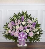 A large, fan-shaped funeral floor basket arrangement featuring lavender stock, pink chrysanthemums, white roses, and white snapdragons, accented with a large lavender satin bow and lush green palm fronds.