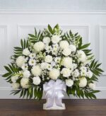 A large, elegant white floral funeral floor basket arrangement featuring roses, mums, daisies, and carnations with lush greenery and a white satin bow.