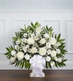 A large, fan-shaped funeral floral arrangement in a white pedestal basket featuring white roses, carnations, chrysanthemums, and snapdragons accented with lush emerald green palm fronds and a large white satin ribbon.