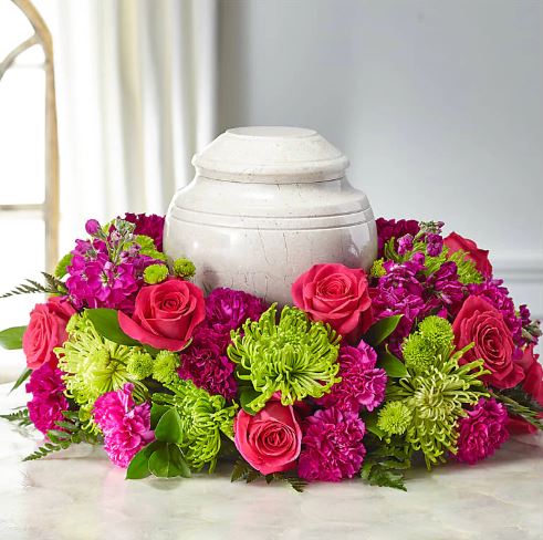 A circular floral wreath arrangement featuring vibrant pink roses, magenta carnations, and bright green spider mums surrounding a polished white marble cremation urn on a light-colored surface.