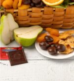 A close-up of a kosher gift basket featuring a sliced pear, Ghirardelli dark chocolate, dried apricots, chocolate-covered nuts, and brittle on a white wooden surface.