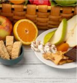 A close-up view of a gift basket arrangement featuring fresh fruits like apples, pears, and oranges alongside sweet treats like biscotti, white chocolate-covered pretzels, peanut brittle, and dark chocolate.