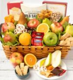 A premium woven gift basket overflowing with assorted fresh fruits including apples, pears, and oranges, along with packaged gourmet treats like Ghirardelli chocolates, smoked salmon, crackers, and nuts.
