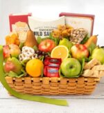 A premium gourmet gift basket woven from light-colored wood, overflowing with a variety of fresh fruits including apples, pears, and oranges, along with packaged snacks such as Ghirardelli chocolates, smoked salmon, crackers, and nuts.