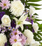 A close-up view of a lavender and white funeral standing spray featuring white roses, lilies, chrysanthemums, and lavender daisies accented with purple statice and greenery.