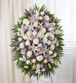 An elegant, large oval-shaped funeral standing spray on a wire easel, featuring a lush mix of white roses, white lilies, white chrysanthemums, lavender daisies, and purple statice, framed by vibrant green palm fronds and emerald leaves against a clean white interior background.