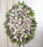 A large, elegant funeral standing spray arrangement featuring a lush mix of white lilies, lavender daisies, white roses, and purple stock flowers, accented with green palm fronds on a wire easel.