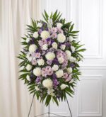 A large, elegant funeral standing spray featuring a blend of lavender, purple, and white flowers including roses, mums, and carnations, arranged with lush green palm fronds on a wire easel.