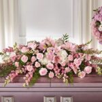 A premium casket spray floral arrangement featuring soft pink lilies, roses, carnations, and white hydrangeas, elegantly draped over a lavender-colored casket.